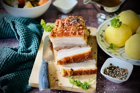 Ein saftiger Schweinebraten mit knuspriger Kruste, auf einem Holzbrett angeschnitten, daneben Klöße und Soße.