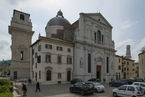La piazza di San Giorgio a Verona, con la chiesa imponente, la torre campanaria e alcune persone che passeggiano tra le auto parcheggiate.
