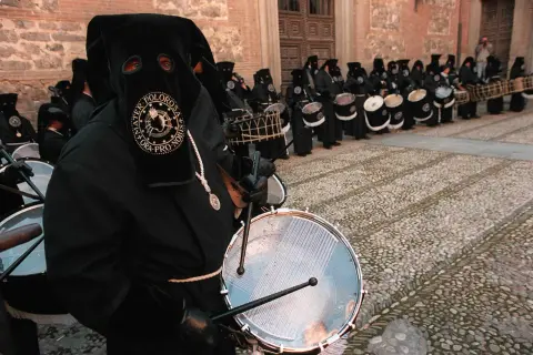 Tambores resuenan en las procesiones de Zaragoza. Nazarenos con túnicas negras y capirotes golpean sus tambores, creando un ritmo solemne.