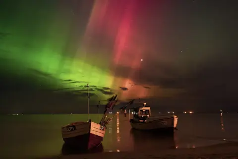 Niezwykła zorza polarna nad Gdańskiem, zielono-czerwone smugi na niebie nad spokojnym morzem i łodziami rybackimi.