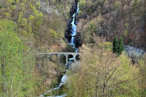 La cascade du Bief de la Ruine dévale une falaise rocheuse, traversée par un pont en arc. La nature verdoyante environnante annonce le printemps.