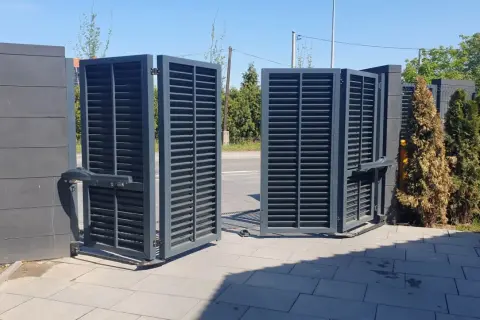 Otwarta brama łamana, szara, z lamelkami, z automatyką, na tle zieleni i nieba.