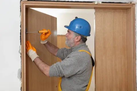 Hombre con casco azul y guantes naranjas ajusta una puerta de contrachapado con un destornillador.
