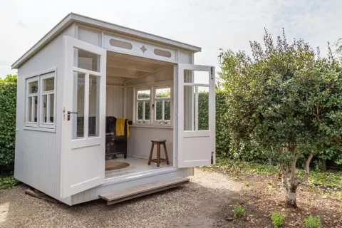 Eine charmante Gartenlaube mit offenen Türen, bereit, mit gemütlichen Möbeln eingerichtet zu werden. Ein Hocker und eine Decke deuten auf eine entspannte Atmosphäre hin.