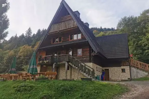 Drewniana bacówka z tarasem i schodami, otoczona zielenią i lasem.