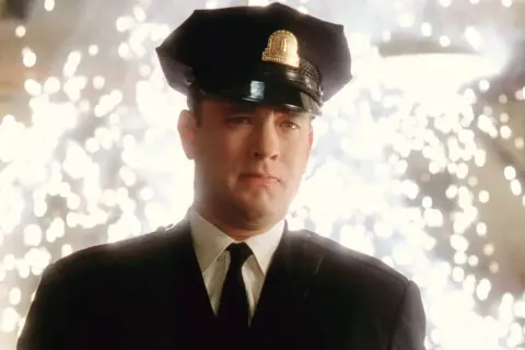 Tom Hanks w mundurze strażnika w jednym z jego filmów. Tło rozświetlone jest fajerwerkami.