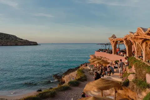 Menschen genießen den Sonnenuntergang am Strand von Ibiza, in der Nähe eines einzigartigen ashram-ähnlichen Gebäudes am Meer.