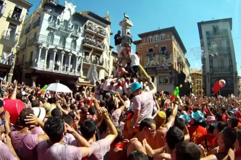 Multitud celebra cuando son vaquillas en Teruel. Gente en una plaza con edificios históricos, algunos subidos a un monumento.
