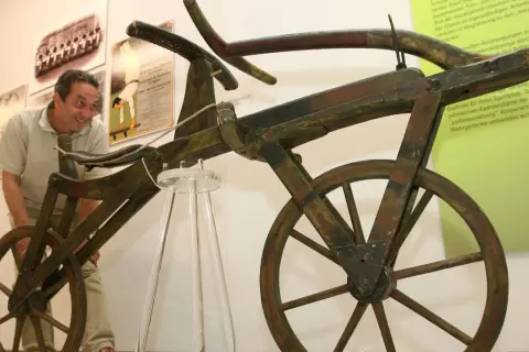 Ein Mann betrachtet das erste Fahrrad der Welt, ein hölzernes Gefährt ohne Pedale, das an ein Laufrad erinnert.