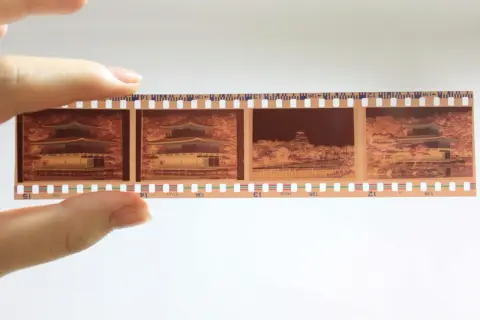En hand håller upp en filmremsa med fyra bilder från en analog kamera. Bilderna visar en vacker byggnad med japansk arkitektur, omgiven av höstfärger.