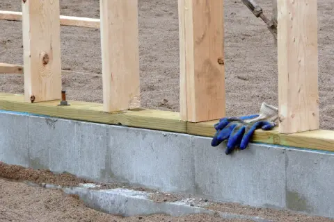 Fundament pod dom drewniany w budowie. Betonowy cokół, drewniane słupki i leżące na belce roboczej niebieskie rękawice budowlane.