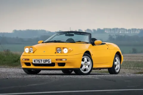 Gul Lotus Elan M100 cabriolet parkerad på grusväg med dimmig bakgrund.