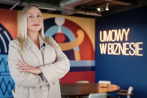 Kobieta w beżowej marynarce z mikrofonem, na tle neonu "UMOWY W BIZNESIE". Czy vider jest bezpieczny?