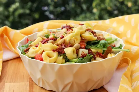 Ein köstlicher tortellini salat mediterran mit sonnengetrockneten Tomaten und Nüssen, serviert in einer weißen Schüssel auf einem Holztisch.