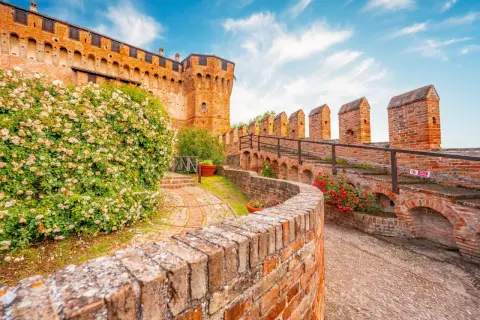 Un sentiero in mattoni conduce al **belvedere ostrense**, con un muro di rose bianche e la rocca medievale sullo sfondo.