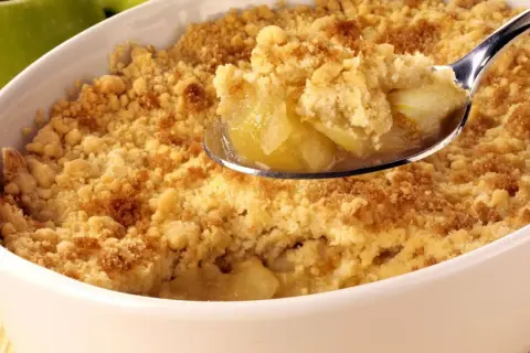 Un délicieux crumble aux pommes, parfait pour suivre la recette de Cyril Lignac. La cuillère en argent révèle des morceaux de pommes fondantes sous une pâte croustillante.
