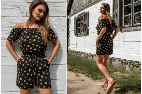 Krótki kombinezon hiszpanka w słoneczniki, idealny na letnie dni. Modelka prezentuje go na tle drewnianej chatki.