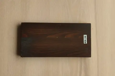 Tavola scura con evidenti venature del legno, su sfondo chiaro con venature simili. Etichetta bianca con scritta "NOCE MEDIO".