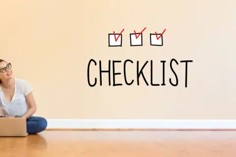 Młoda kobieta z laptopem siedzi na podłodze. Na ścianie nad nią widnieje napis "CHECKLIST" z trzema zaznaczonymi polami.