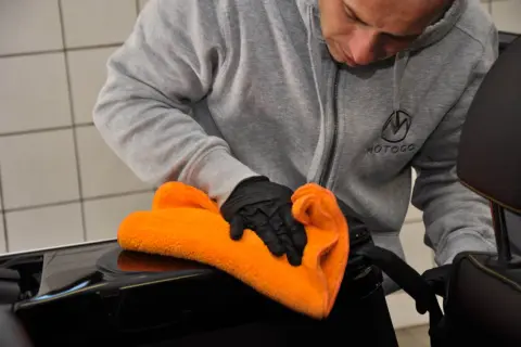 Mężczyzna w rękawiczkach czyści czarną powierzchnię pomarańczową mikrofibrą. Używa adbl apc pro do pielęgnacji wnętrza auta.