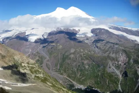 Elbrus, najwyższy szczyt Kaukazu, w chmurach, z lodowcami i zielonymi dolinami poniżej.