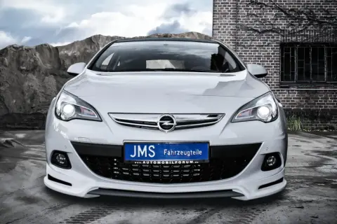 Biały Opel Astra J GTC z tuningiem JMS, z tablicą rejestracyjną "JMS Fahrzeugteile", na tle kamieniołomu i ceglanego budynku.