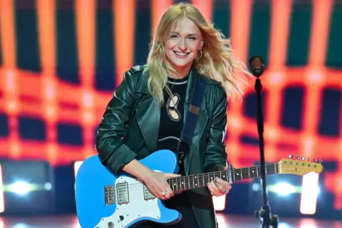 Uśmiechnięta polska piosenkarka z blond włosami gra na niebieskiej gitarze elektrycznej na tle pomarańczowych świateł.
