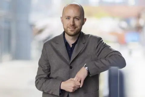 Viktor Barth Kron, en man med kortklippt hår och skägg, bär en brun kavaj och en mörkblå skjorta. Han lutar sig mot ett räcke utomhus.