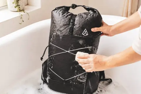Person reinigt einen schwarzen Rucksack in einer Badewanne mit Schaum. Der Rucksack wird mit einem Schwamm abgerieben, um ihn zu waschen.