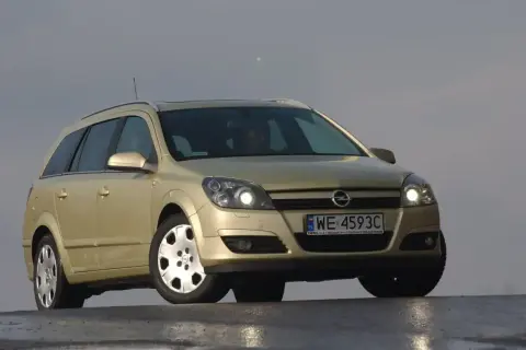 Złoty Opel Astra H kombi z włączonymi światłami. Przód samochodu z wyraźnie widocznymi żarówkami.