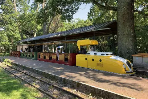 Die gelbe Lokomotive der Schlossbahn Karlsruhe steht bereit. Fahrgäste sitzen in den roten und grünen Waggons unter einem schattigen Dach.