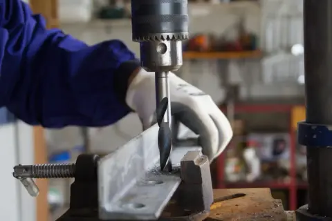 Ręka w rękawicy obsługuje wiertarkę stołową, wiercąc w metalowym profilu. Widać części do wiertarki stołowej i wióry metalowe.
