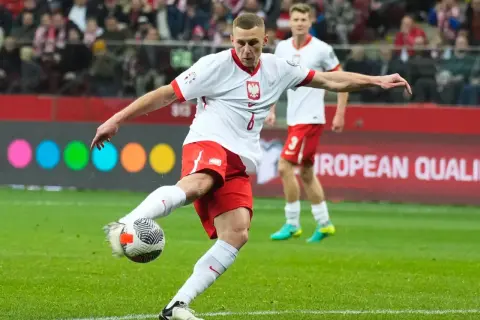 Piłkarz w białej koszulce z orłem i czerwonych spodenkach kopie piłkę na boisku.