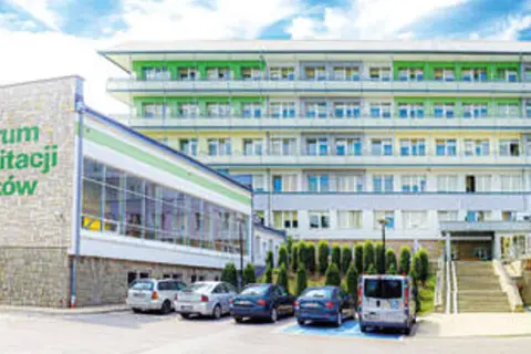 Sanatorium z rehabilitacją kręgosłupa dla rolników. Nowoczesny budynek z zielonymi elementami fasady, otoczony drzewami i parkingiem.