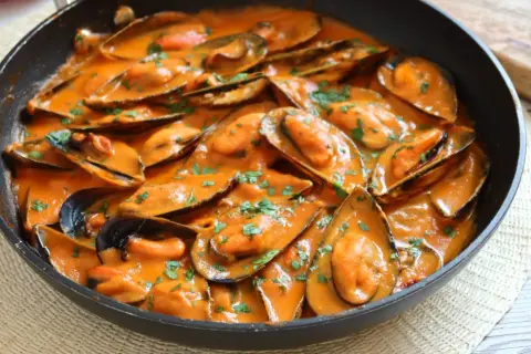 Mejillones con tomate en salsa cremosa, espolvoreados con perejil fresco, servidos en una sartén negra.