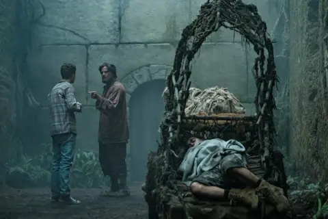 Zwei Männer diskutieren in einer mysteriösen, moosbewachsenen Kammer. Im Vordergrund schläft eine Person in einem rustikalen Bett. Szene aus "Der Greif Staffel 2".