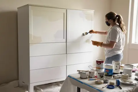 Kobieta maluje białe mdf meble na jasny kolor. Na stole stoją puszki z farbą i taśmy malarskie.