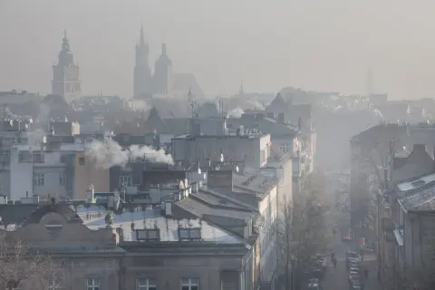 Widok na miasto w smogu z dymiącymi kominami i zarysami wież kościelnych w tle.