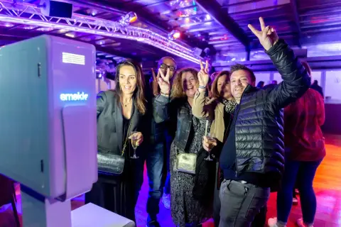 Des amis s'amusent devant un photobooth, faisant des signes de paix. L'ambiance est festive, éclairée par des lumières violettes.