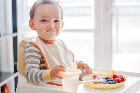 Kleiner Junge mit Heidelbeeren und Joghurt im Gesicht, der an seinem Becher nippt.
