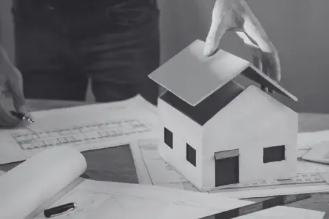 Model domu na papierach z planami. Ręka kładzie dach, symbolizując sprzeciw sąsiada na budowę lub trudności w uzyskaniu pozwolenia.