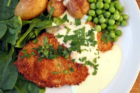 Krispig schnitzel med krämig sås till schnitzel, serverad med små potatisar, gröna ärtor och färsk spenat.