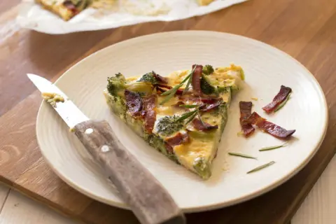 Une part de quiche sans pâte aux brocolis et lardons, garnie de romarin, sur une assiette avec un couteau.