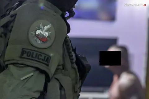 Funkcjonariusz CBŚ w kamizelce z napisem POLICJA i godłem Polski. W tle mężczyzna z zamaskowaną twarzą.