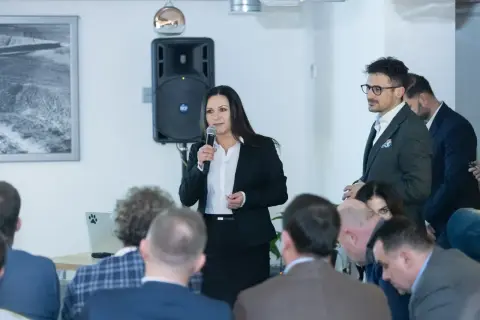 Kobieta w czarnej marynarce przemawia do grupy ludzi, trzymając mikrofon.