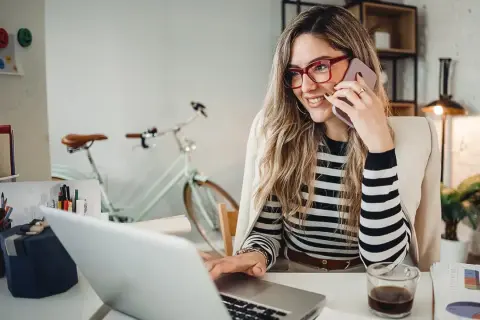Frau am Telefon, lächelnd, am Laptop arbeitend. Hinter ihr ein Fahrrad. Perfekt für den **iu semesterbeginn**-Stress.