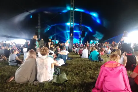 Widownia siedzi na trawie pod namiotem, czekając na koncert. Niebieskie światła oświetlają scenę, tworząc atmosferę festiwalu Open'er.