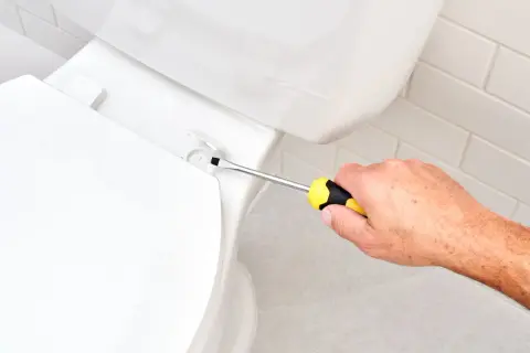 A hand uses a screwdriver to tighten a bolt, demonstrating how to install a toilet seat.