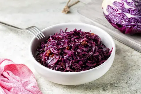 Un bol de chou rouge recette, garni de raisins secs, prêt à être dégusté. Une moitié de chou rouge est visible sur une planche à découper en arrière-plan.