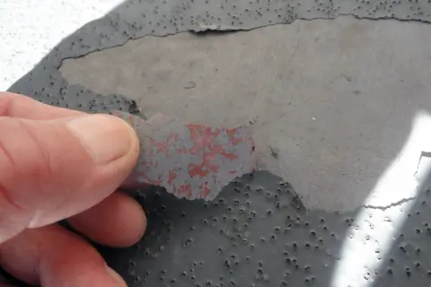 Dedo retirando capa desprendida de pintura gris sobre hormigón con picaduras, indicando necesidad de reparación de hormigón.
